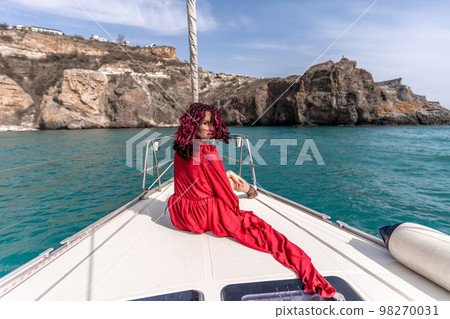 Attractive middle-aged woman in a red dress on a yacht on a summer day. Luxury summer adventure, outdoor activities. 98270031