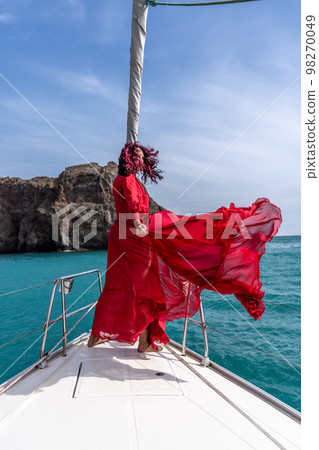 Attractive middle-aged woman in a red dress on a yacht on a summer day. Luxury summer adventure, outdoor activities. 98270049
