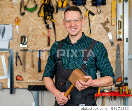 Portrait of a carpenter in goggles and overalls holding a wooden hammer in the workshop against the background of a wall with tools. 98271383