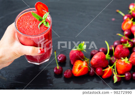 Woman hand holds berries smoothie with strawberries and cherries on black table. Selective focus Woman hand holds berries smoothie with strawberries and cherries on black table. Selective focus 98272990