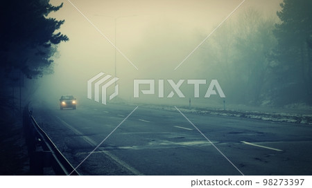 Car traveling on the foggy road with headlights or headlamps on. Low visibility - Dangerous driving of cars in winter in bad weather. 98273397