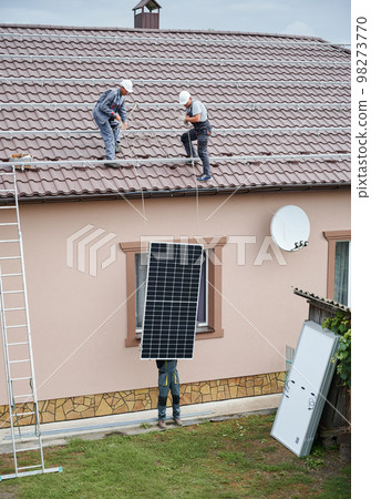Men technicians lifting up photovoltaic solar modul on roof of house. Engineers in helmets building solar panel system outdoors. Concept of alternative and renewable energy. 98273770