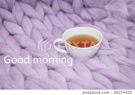 The concept of coziness and comfort is a pink-lavender knitted blanket with a white cup of tea on it. Up close Good morning inscriptions. The concept of coziness and comfort is a pink-lavender knitted blanket with a white cup of tea on it. Up close Good morning inscriptions. 98274420