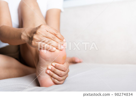 Foot pain, Asian woman sitting on sofa feeling pain in her foot at home, female suffering from feet ache use hand massage relax muscle from soles in home interior, Healthcare and podiatry medical 98274834