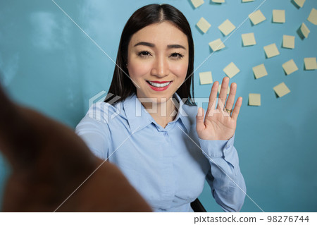 Businesswoman preparing the camera phone for a video conference with business partners, smiling and waving at the camera. Female project manager on virtual call with work team. 98276744