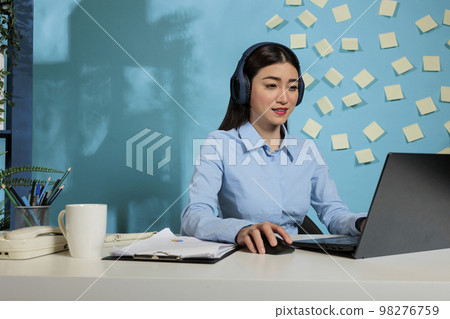 Professional woman working on a laptop with headphones listening to online training in the office. Female corporate worker concentrating at workstation with wireless headset. 98276759
