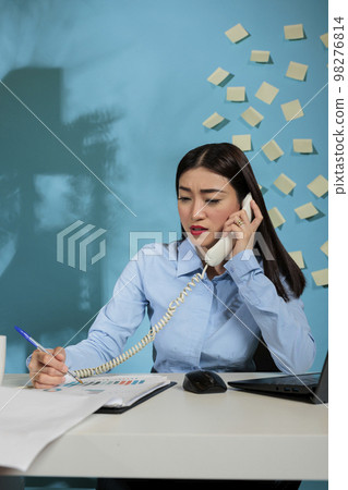 Busy asian woman in startup office talking on landline phone looking at papers with sales charts on desk. Business analyst talks with work team member explaining report results. Busy asian woman in startup office talking on landline phone looking at papers with sales charts on desk. Business analyst talks with work team member explaining report results. 98276814