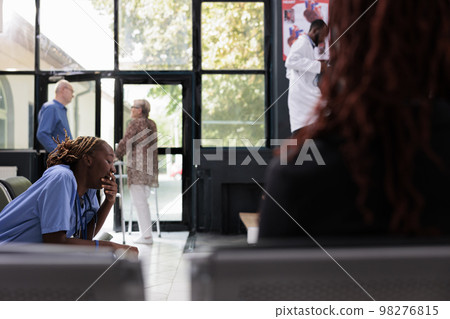 Selective focus of tired exhausted medical assistant sitting on chair in hospital waiting area having a break before start examination. African american nurse working at patient health care treatment Selective focus of tired exhausted medical assistant sitting on chair in hospital waiting area having a break before start examination. African american nurse working at patient health care treatment 98276815