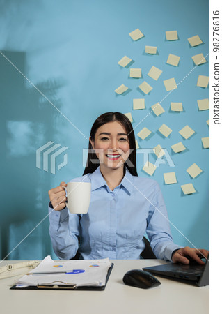 Optimistic woman sitting at workstation drinking a cup of coffee while working with a laptop. Casual startup employee relaxing at work with statistics in a business workplace. Optimistic woman sitting at workstation drinking a cup of coffee while working with a laptop. Casual startup employee relaxing at work with statistics in a business workplace. 98276816