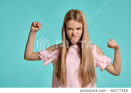 Studio portrait of a beautiful girl blonde teenager in a pink t-shirt posing over a blue background. 98277486