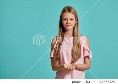 Studio portrait of a beautiful girl blonde teenager in a pink t-shirt posing over a blue background. 98277520