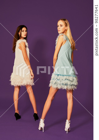 Professional photo of two young women, blonde and brunette, in cocktail dresses posing on purlpe background Professional photo of two young women, blonde and brunette, in cocktail dresses posing on purlpe background 98277641