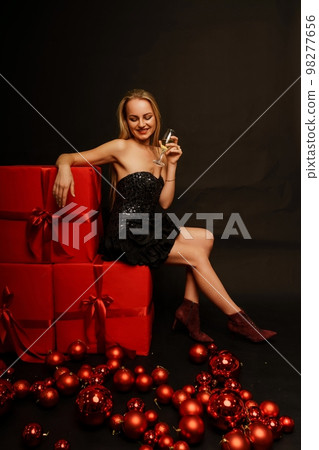 Woman gifts christmas. Beautiful sexy young blonde in a black dress sits on three big red gifts on a black background. Christmas photo, gifts Woman gifts christmas. Beautiful sexy young blonde in a black dress sits on three big red gifts on a black background. Christmas photo, gifts 98277656