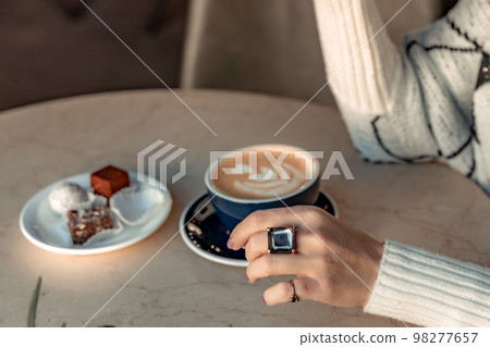 A cup of fresh cappuccino coffee in the hands of a woman on a fashionable background of a white marble table, next to a plate with sweets. Coffee addiction A cup of fresh cappuccino coffee in the hands of a woman on a fashionable background of a white marble table, next to a plate with sweets. Coffee addiction 98277657