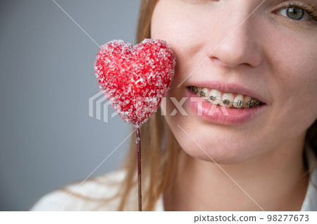 Cute woman with braces on her teeth holds a candy in the form of a heart on white background Cute woman with braces on her teeth holds a candy in the form of a heart on white background 98277673