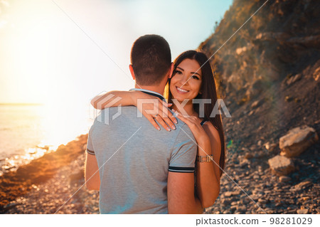 A smiling woman embraces a man. The view from the back. A young couple of lovers pose against the background of the beach, sea and sunset. The concept of Valentine's day A smiling woman embraces a man. The view from the back. A young couple of lovers pose against the background of the beach, sea and sunset. The concept of Valentine's day 98281029