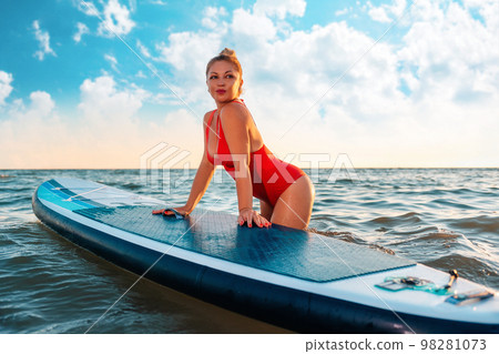 A sexy adult tanned woman in red swimsuit poses with sup board at the sea. The concept of Surfing 98281073
