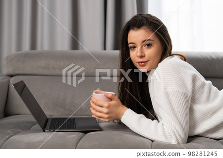 Portrait of a beautiful young girl lying on a sofa with a cup of tea and a laptop. Portrait of a beautiful young girl lying on a sofa with a cup of tea and a laptop. 98281245