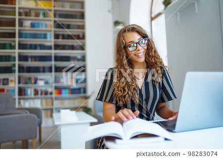 Young woman sitting with a book, laptop in the library and taking learning notes. Concept of education. Young woman sitting with a book, laptop in the library and taking learning notes. Concept of education. 98283074