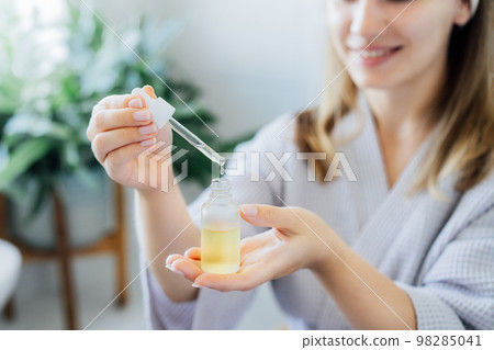 Close up young woman in bathrobe holding mockup bottle and dropper with hyaluronic acid serum in her hands. Skin hydrating. Cosmetic spa procedures. Beauty self-care at home. Selective focus. 98285041