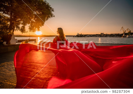 Sunrise red dress. A woman in a long red dress against the backdrop of sunrise, bright golden light of the sun's rays. The concept of femininity, harmony. 98287304