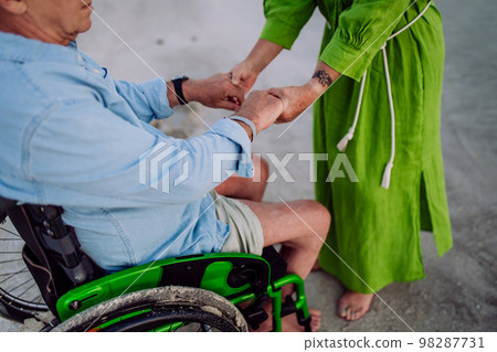 Close-up of senior woman holding hands her husband on wheelchair, enjoying vacations together. Close-up of senior woman holding hands her husband on wheelchair, enjoying vacations together. 98287731