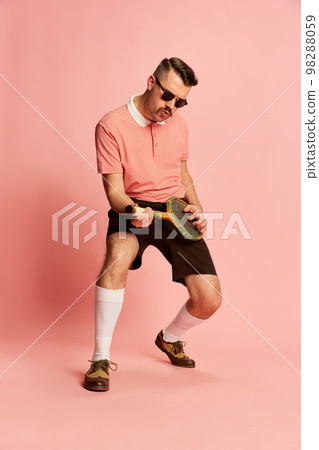 Portraits of handsome charismatic man in stylish clothes posing, playing tennis racket like guitar over pink studio background. Concept of fashion, sport, lifestyle 98288059