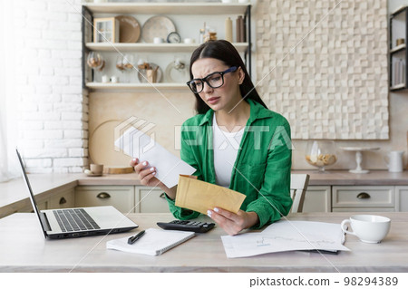 Worried young woman working in the kitchen at home with a laptop. She is holding an envelope with a letter in her hands, she received bad news, an account, debts, divorce papers. 98294389