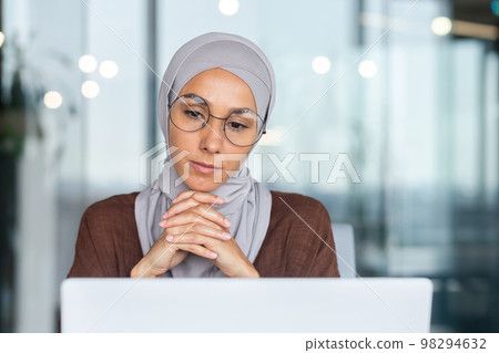 Serious bored businesswoman inside office, muslim woman in hijab thinking while sitting at workplace with laptop, woman at work thinking about decisions. Serious bored businesswoman inside office, muslim woman in hijab thinking while sitting at workplace with laptop, woman at work thinking about decisions. 98294632
