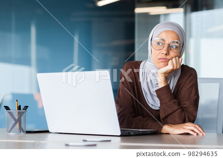 Serious bored businesswoman inside office, muslim woman in hijab thinking while sitting at workplace with laptop, woman at work thinking about decisions. Serious bored businesswoman inside office, muslim woman in hijab thinking while sitting at workplace with laptop, woman at work thinking about decisions. 98294635