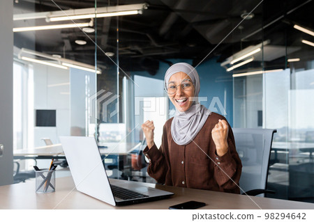 Portrait of successful businesswoman in hijab, muslim woman working inside office using laptop at work looking at camera and smiling happy, female worker celebrating victory good achievement results 98294642