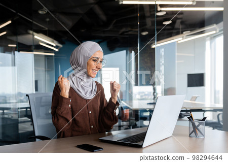 Successful businesswoman in hijab celebrating victory and successful achievement of results, woman looking at laptop screen and holding hands up in win and triumph gesture. 98294644