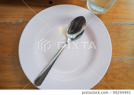 An empty white plate with a spoon is placed on a wooden table. An empty white plate with a spoon is placed on a wooden table. 98294991