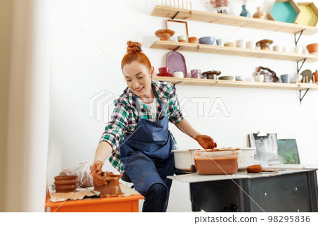 Cheerful female sculptor while she making clay pot on pottery wheel. Idea of small business and entrepreneurship. Home hobby, entertainment and leisure. Art studio Cheerful female sculptor while she making clay pot on pottery wheel. Idea of small business and entrepreneurship. Home hobby, entertainment and leisure. Art studio 98295836