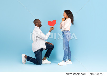 Romantic african american man on his knees giving red heart shaped card to excited black woman, side view Romantic african american man on his knees giving red heart shaped card to excited black woman, side view 98298756