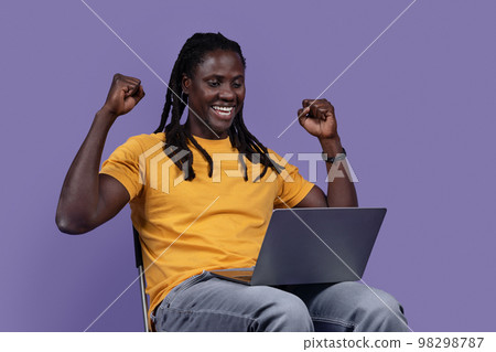 Happy black man sitting on chair, using laptop and gesturing 98298787