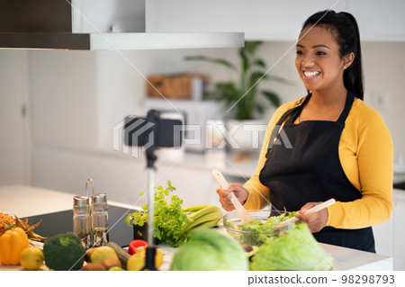 Happy young african american woman in apron prepare salad at table with fresh vegetables, shoot video on phone 98298793