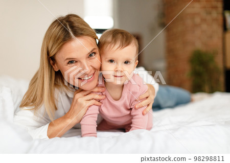 Happy motherhood. Mommy playing with adorable toddler baby crawling on bed, smiling at camera, bedroom interior 98298811