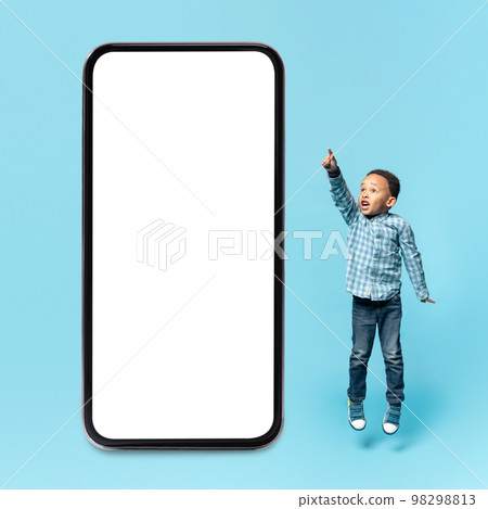 Educational kids mobile app. Surprised black little boy pointing aside and jumping near big smartphone with blank screen Educational kids mobile app. Surprised black little boy pointing aside and jumping near big smartphone with blank screen 98298813