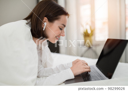 Cheerful pretty woman using laptop and earpods in bed, mockup 98299026