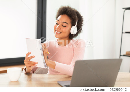 Smiling dreamy pensive young mixed race female manager in wireless headphones makes notes 98299042