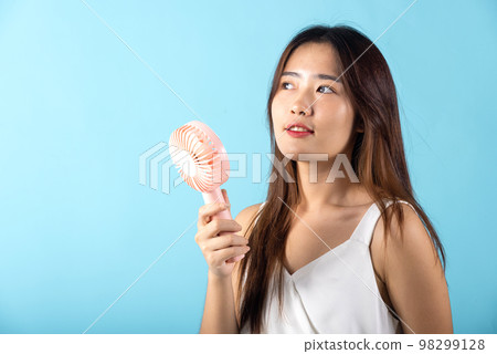 Asian beauty young woman holding pink portable electric mini fan near her face studio shot isolated on blue background, Female hand hold small plastic fan handheld she enjoying cool wind blowing 98299128