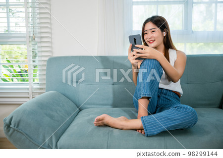 Social media networks. Happy beautiful female relax and shopping online with smartphone, Smiling Asian woman sitting on sofa reading news on mobile smart phone at living room at home Social media networks. Happy beautiful female relax and shopping online with smartphone, Smiling Asian woman sitting on sofa reading news on mobile smart phone at living room at home 98299144