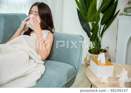 Asian young woman she sick blowing nose sneezing in tissue at home, fever caught cold, Sick female sitting under blanket on sofa and sneeze with tissue paper in living room, medicine healthcare Asian young woman she sick blowing nose sneezing in tissue at home, fever caught cold, Sick female sitting under blanket on sofa and sneeze with tissue paper in living room, medicine healthcare 98299147