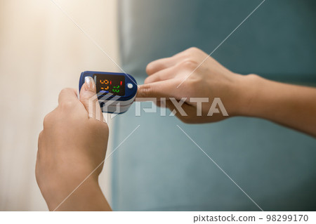 Young woman using pulse oximeter to monitor blood oxygen levels and pulse rate on finger under sofa in living room at home, Self healthcare with coronavirus pandemic, medical healthcare concept 98299170