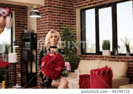 Woman holding valentines day expensive gift, looking at camera with calm lovely facial expression. Beautiful girl in black dress standing in living room filled with presents and red roses bouquet Woman holding valentines day expensive gift, looking at camera with calm lovely facial expression. Beautiful girl in black dress standing in living room filled with presents and red roses bouquet 98299726