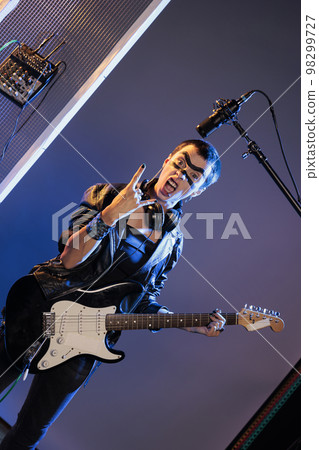 Punk rocker doing rock sign and playing guitar, feeling ecstatic and doing solo concert in studio over background. Female singer showing rock and roll symbol at live show performance. Punk rocker doing rock sign and playing guitar, feeling ecstatic and doing solo concert in studio over background. Female singer showing rock and roll symbol at live show performance. 98299727
