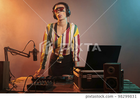 Flirty disc jockey acting silly with tongue out, fooling around in studio with lights. Playful funky woman mixing techno music with mixer or turntables, being confident and laid back. 98299831