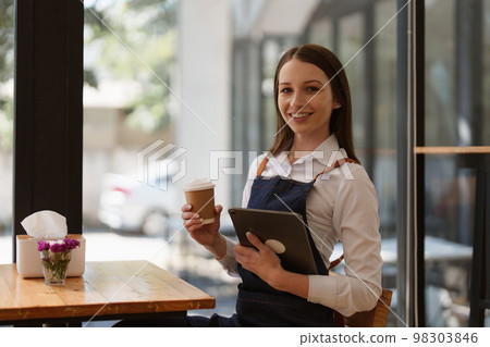Young Female manager in restaurant with tablet. Woman coffee shop owner with open sign. Small business concept Young Female manager in restaurant with tablet. Woman coffee shop owner with open sign. Small business concept 98303846
