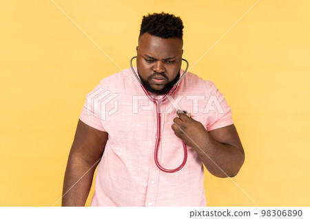 Portrait of concentrated bearded man wearing pink shirt standing with stethoscope, doing self examining of his heart, medicine concept. Indoor studio shot isolated on yellow background. 98306890
